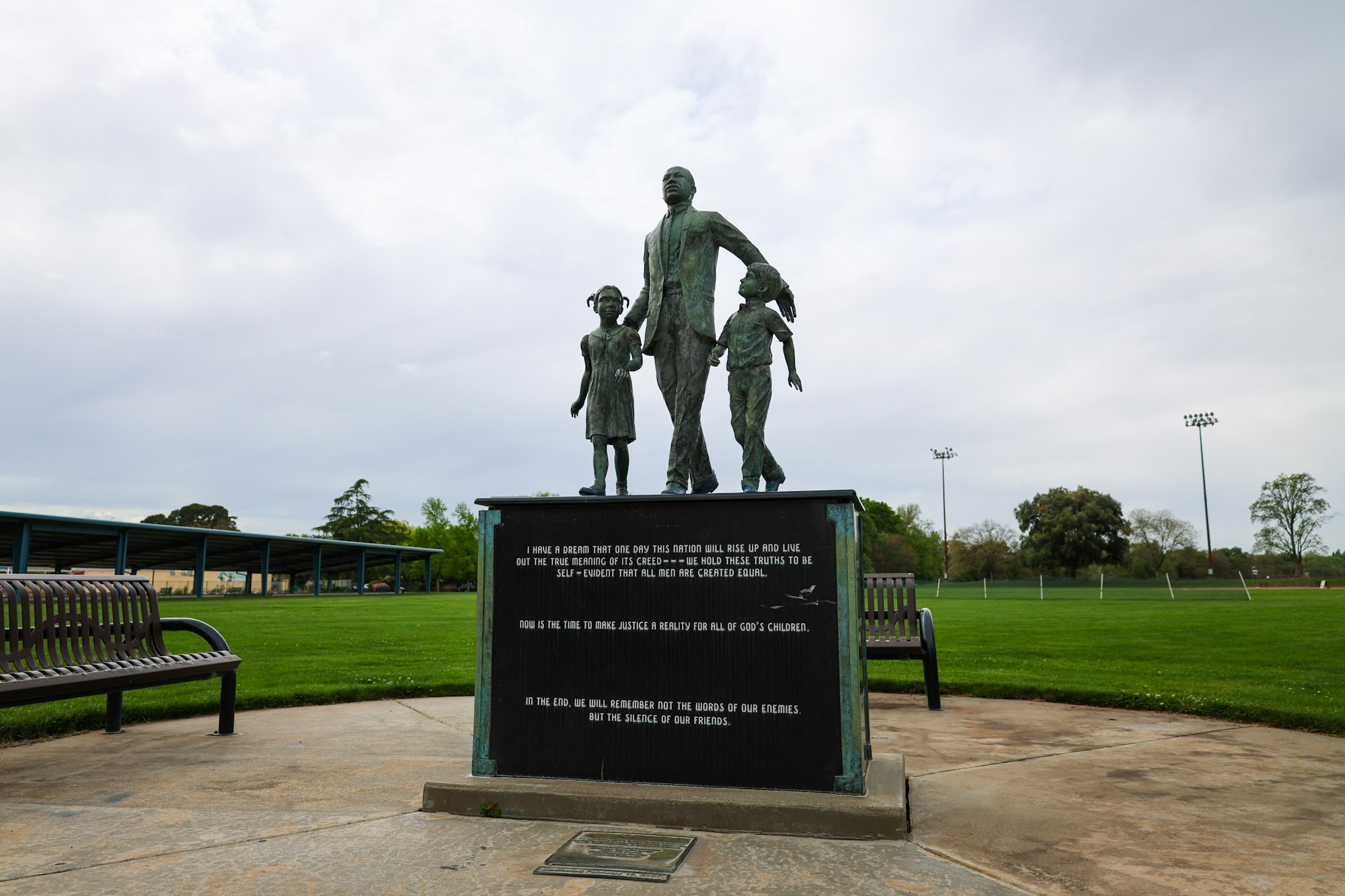 Statue of a man with two children and a plaque with inspirational quotes, set in a park with a sports field in the background.