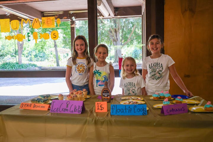 Four kids at a table selling ocean brownies, ice cream cake pops, floatie cookies, and cupcakes at a "Summer Sweets" stand.