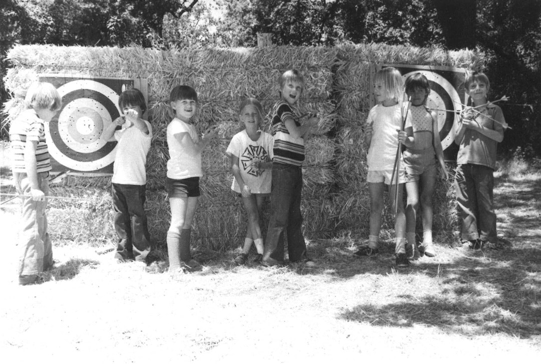 Kids with bows and arrows posing in front of hay bales with archery targets.