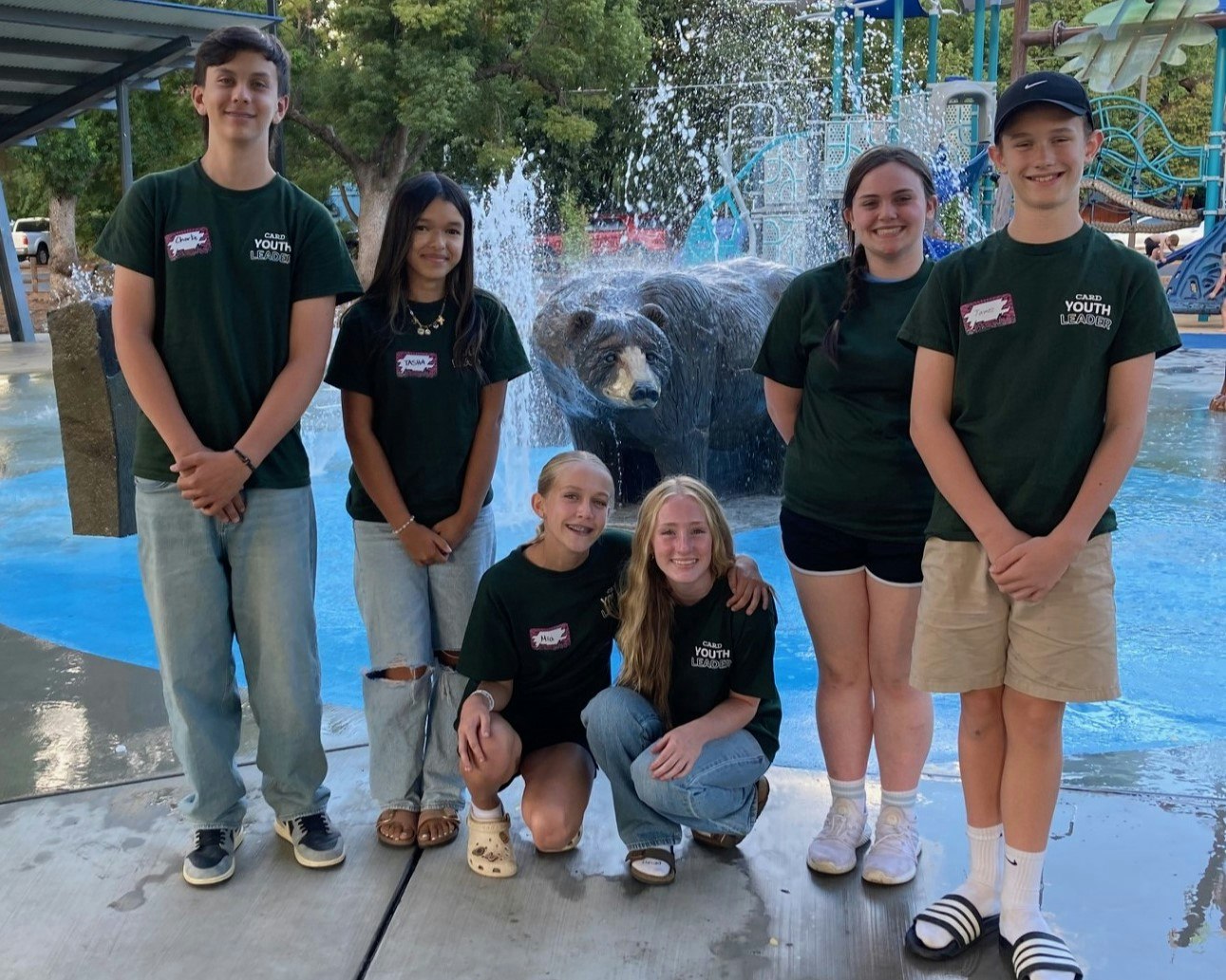 Six youth leaders in green shirts pose together outside near a fountain and a bear statue.