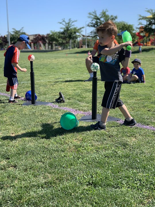 Kids are playing in a park, participating in a baseball practice with a tee, while others watch nearby in a sunny, green setting.