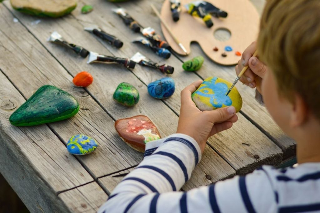 A child painting colorful designs on rocks with paint tubes and a wooden palette on a wooden table.
