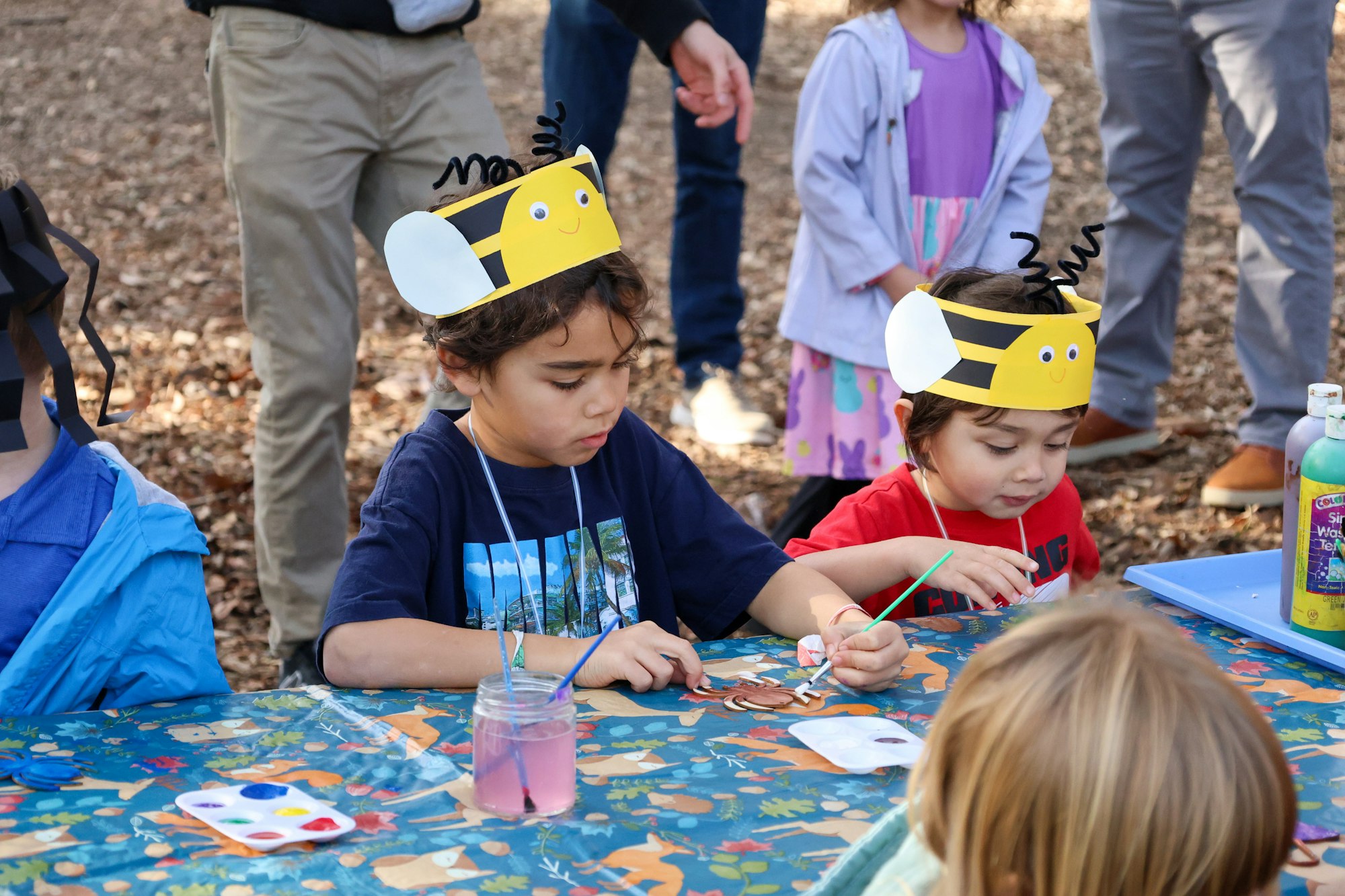 Kids wearing bee hats craft and paint at a table outdoors, with colorful supplies and playful decorations around them.