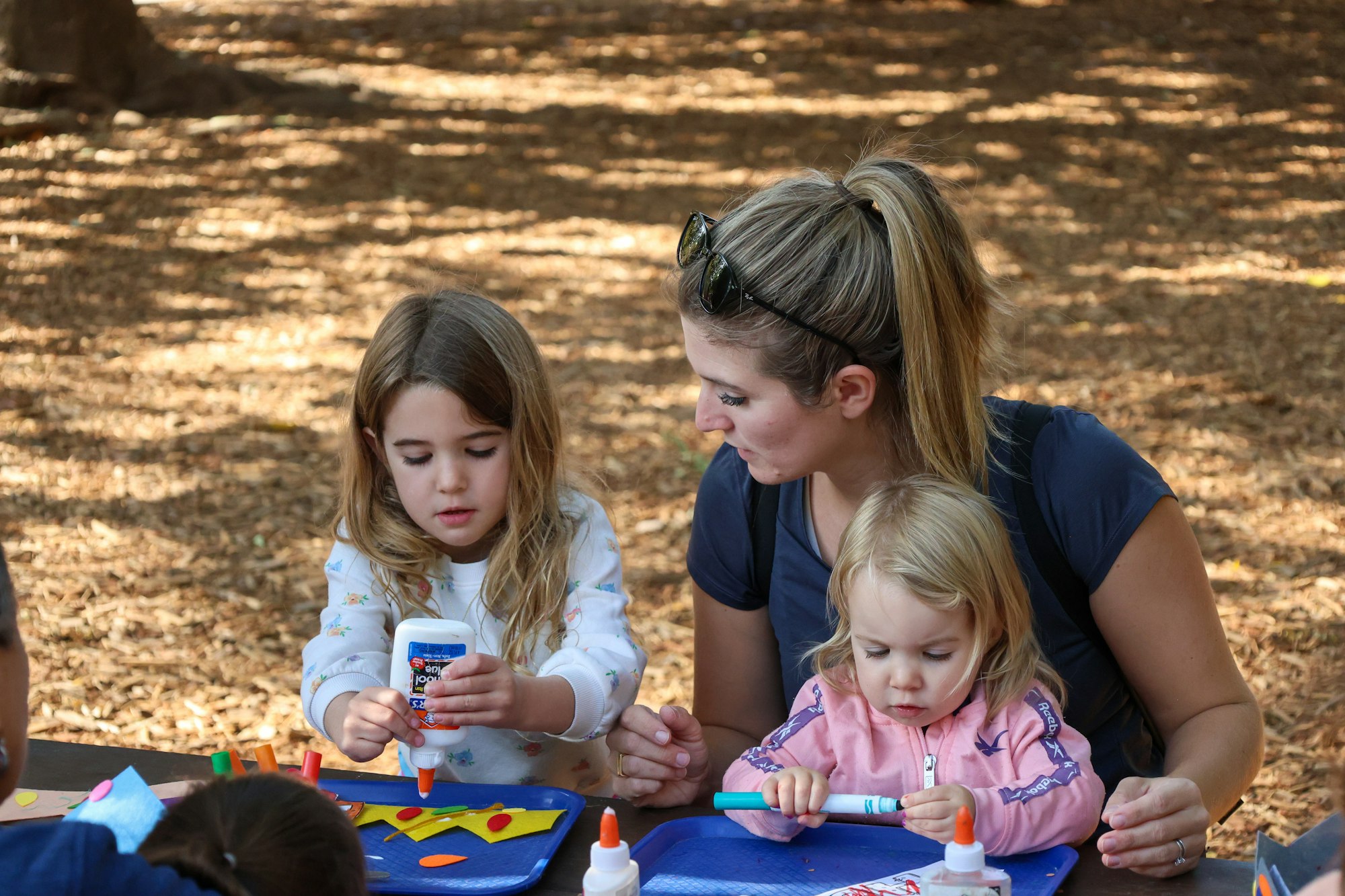 A woman and two young girls are engaged in a craft activity outdoors, using glue and markers on colorful materials.