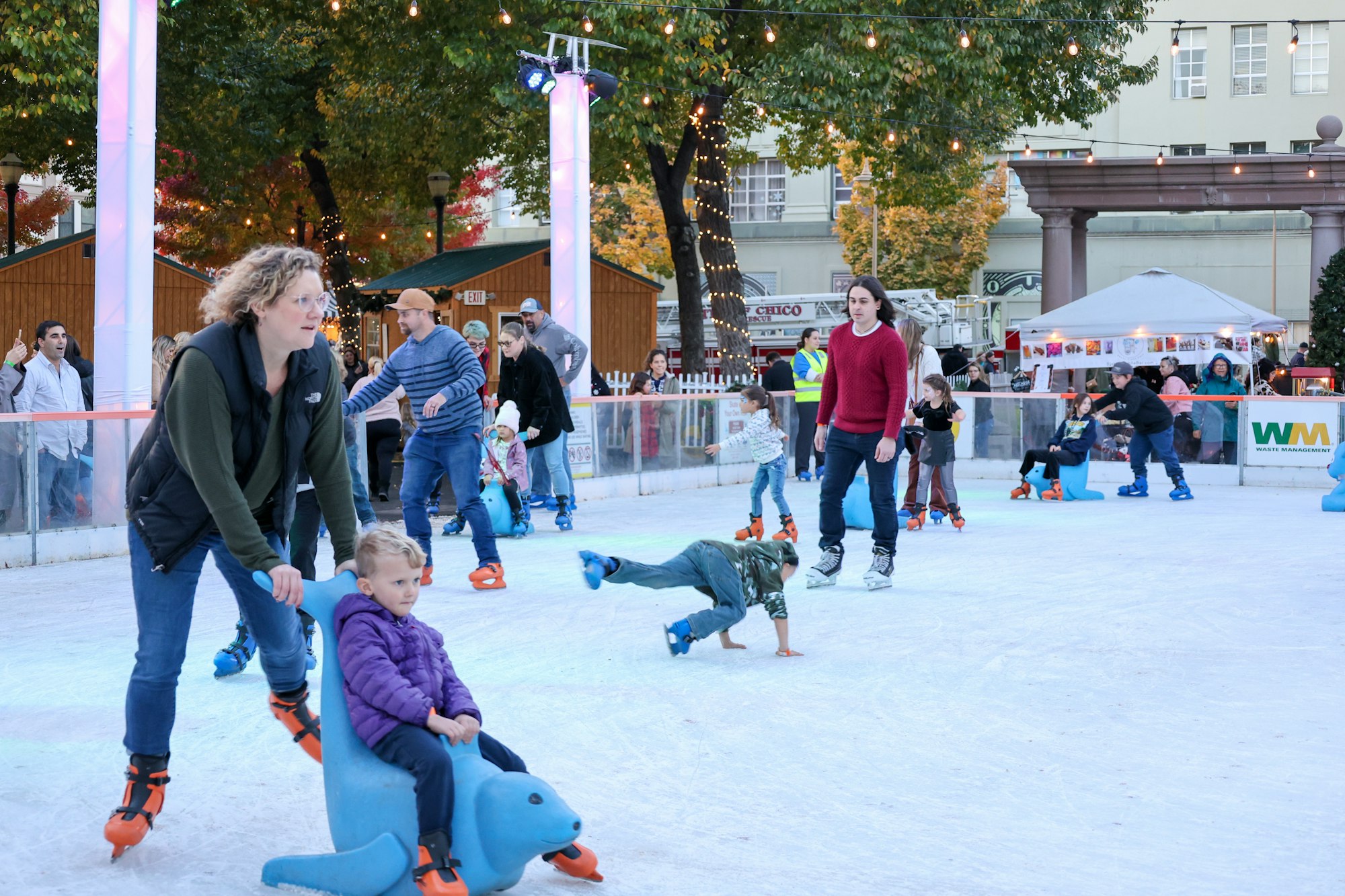 An outdoor ice skating rink with people skating, including a child on a push-along seal aid.