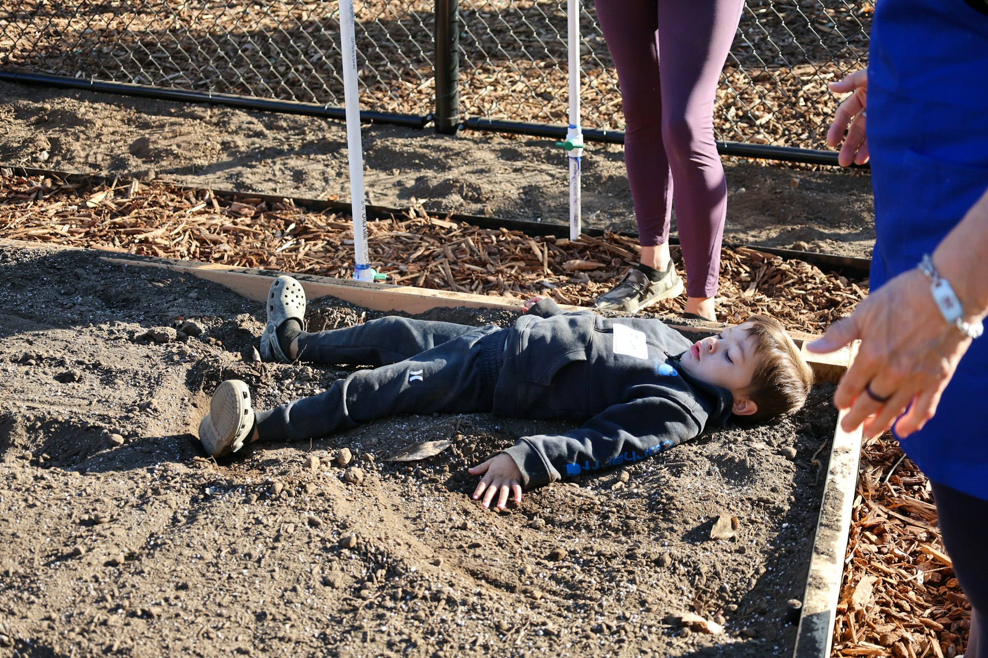A child lies on the dirt surrounded by adults, near a fence and wood chips.