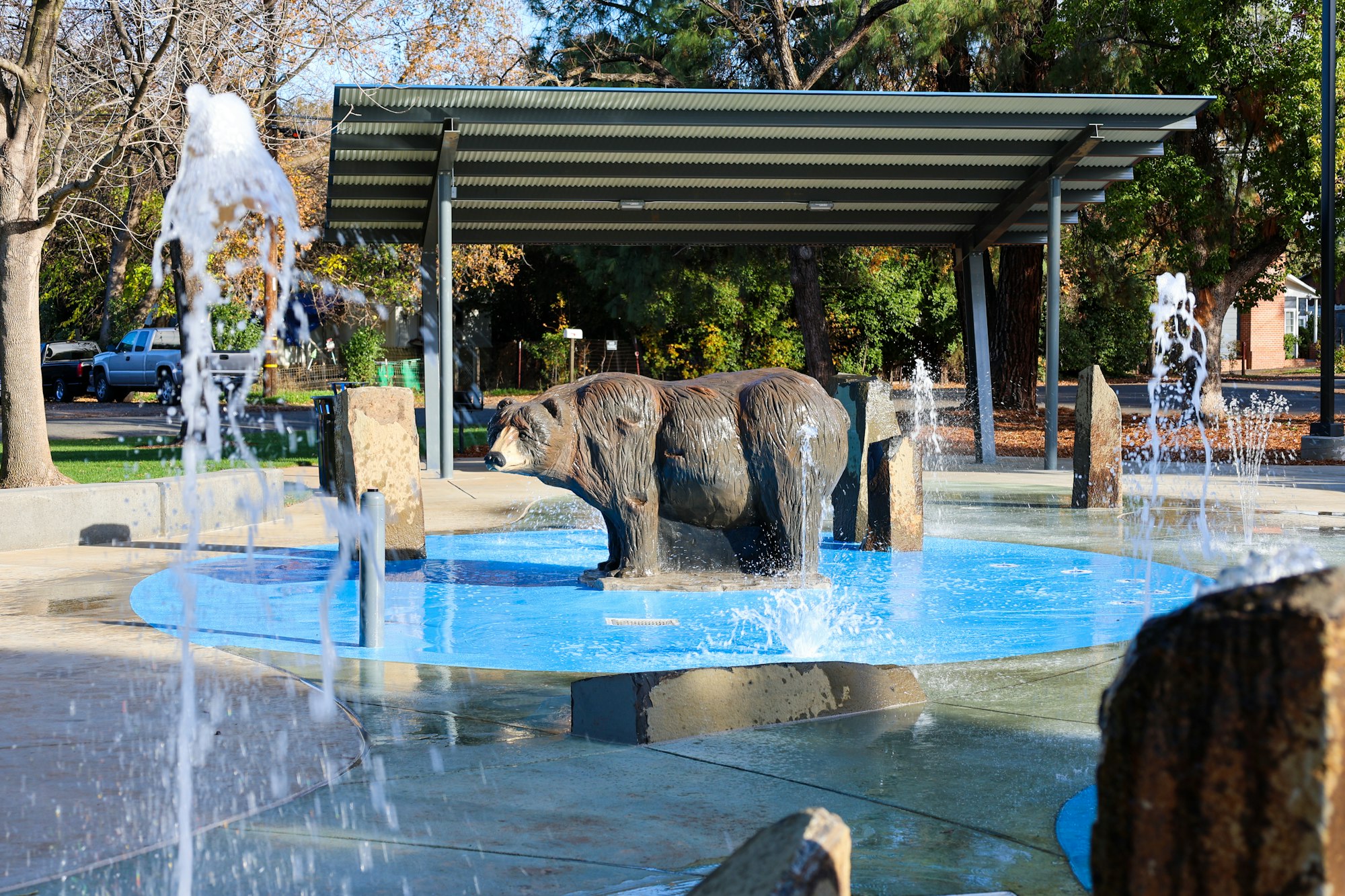 A bear sculpture in a fountain with water jets, surrounded by trees and a shelter in the background.