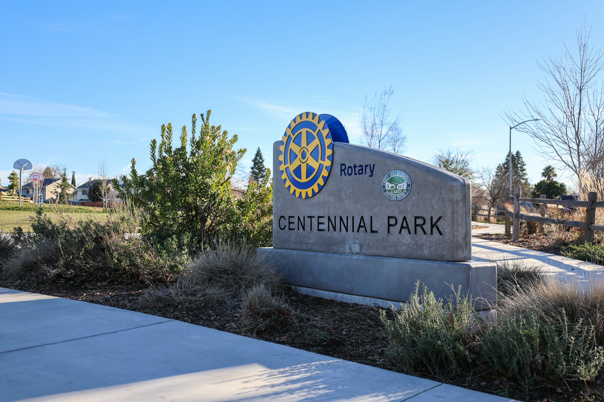 Centennial Park entrance sign with Rotary logo, surrounded by greenery and a clear sky.