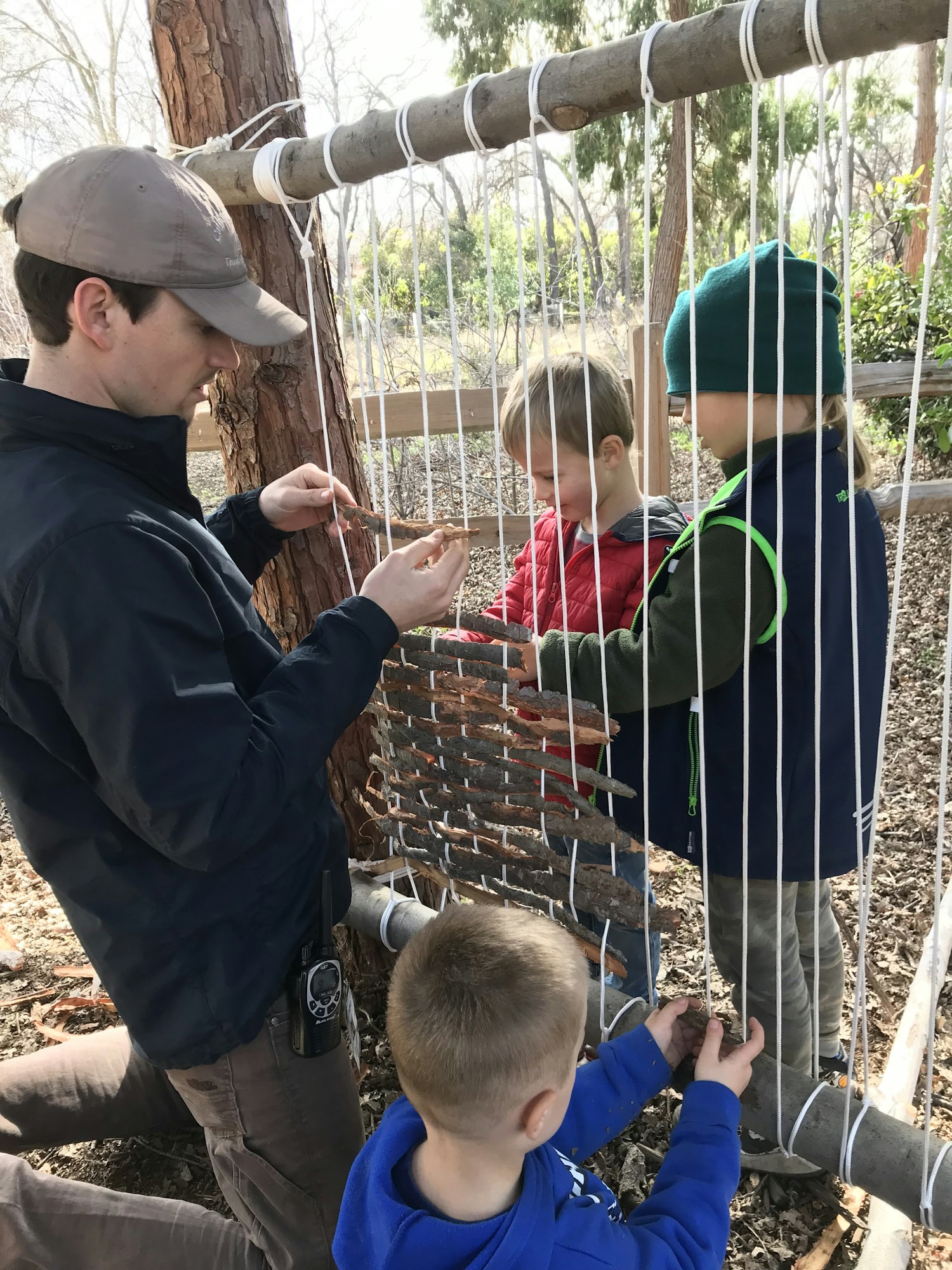 Children and an adult weaving sticks into a vertical loom made of white ropes outdoors.