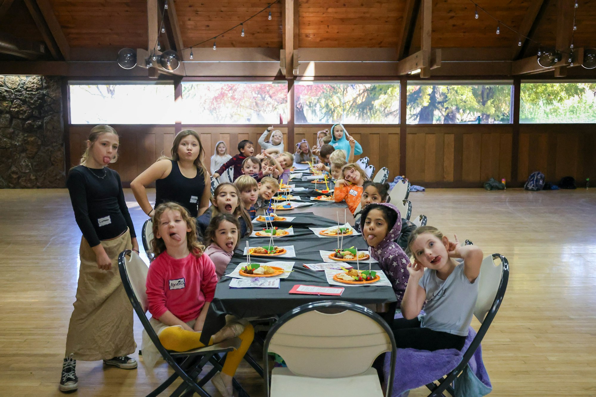A group of children at a long table, making funny faces with plates of food in front of them, inside a wood-paneled room.