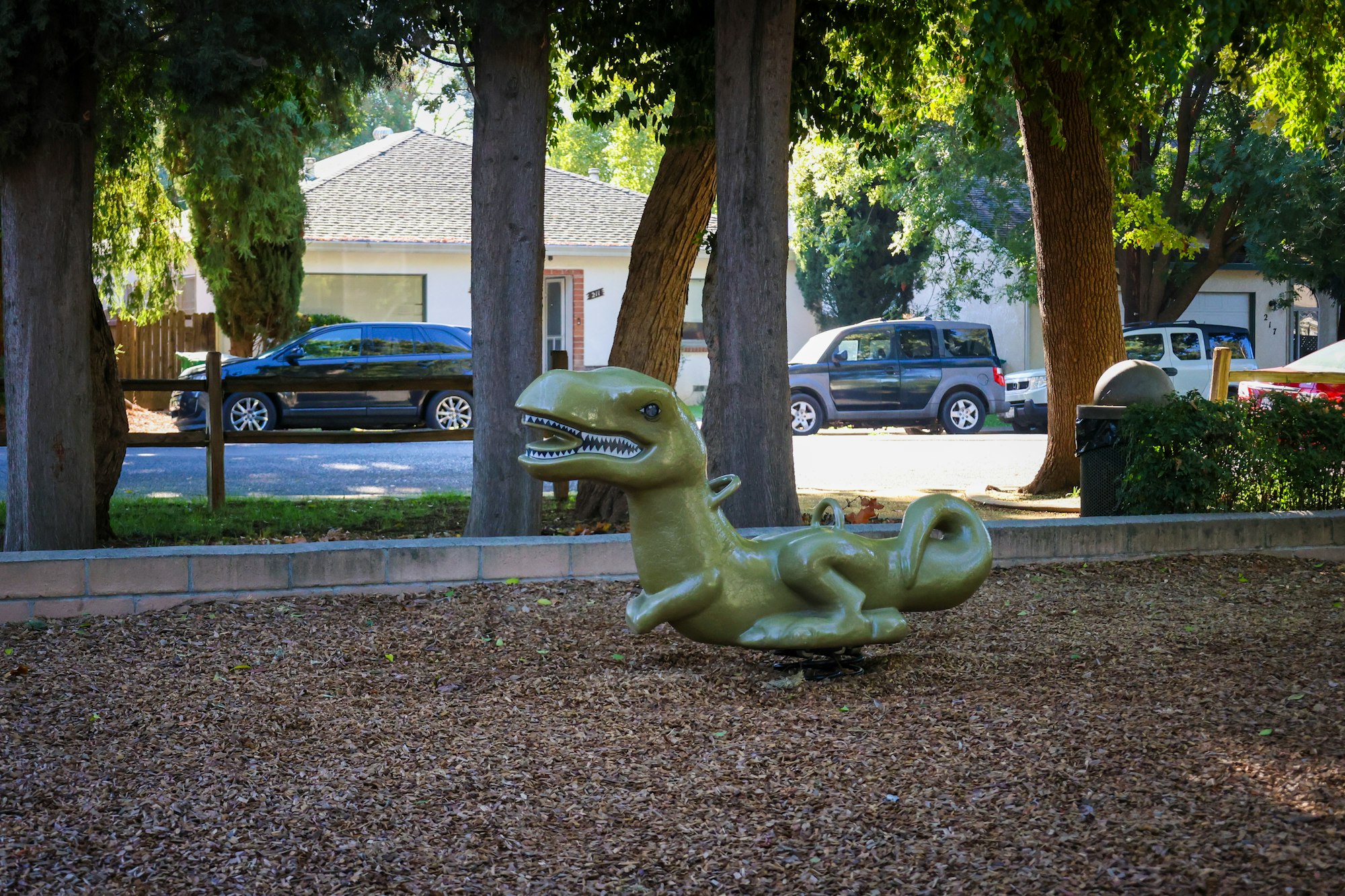 A green dinosaur-shaped spring rider in a park surrounded by trees and parked cars.