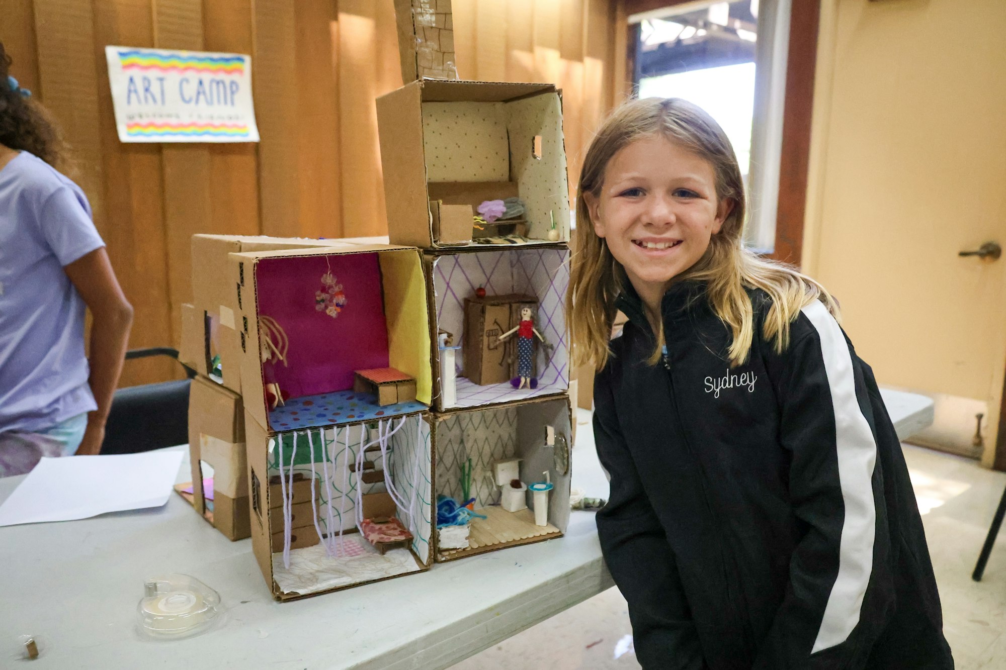 A child smiles beside a homemade cardboard dollhouse at an art camp.