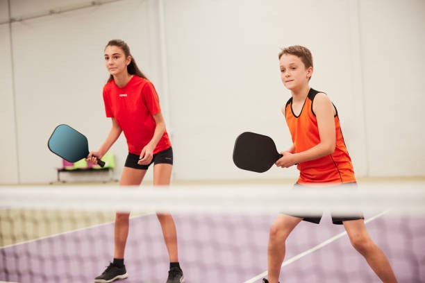 Two kids are preparing to play a game, each holding a paddle, in an indoor tennis or pickleball court.