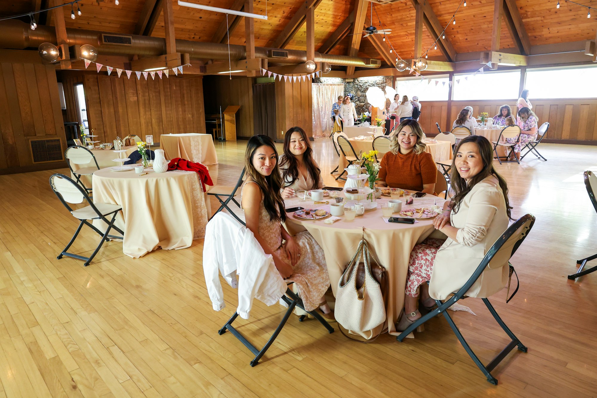 A group of people sitting at round tables in a wooden hall, enjoying a gathering with food and decorations.