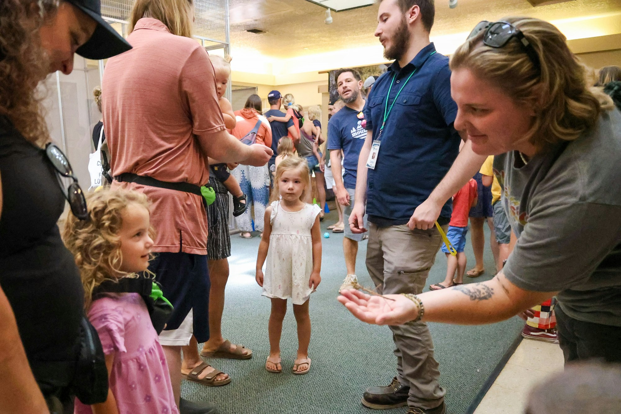 A group of people, including children, gather indoors. An adult extends their hand, engaging with the children.