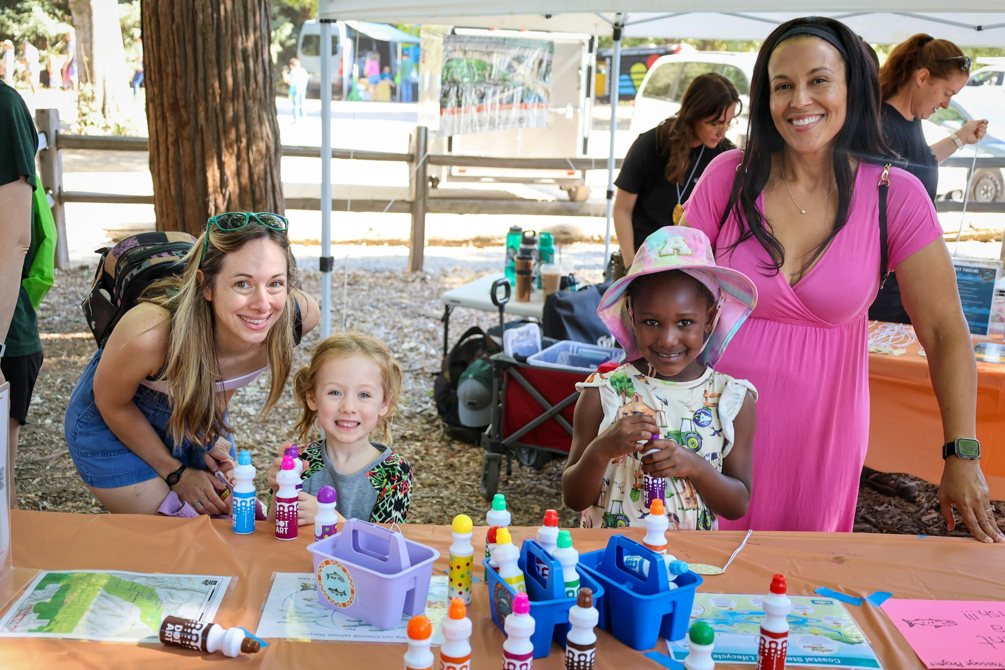 A group of people, including children, enjoying a craft activity at an outdoor event with art supplies on a table.