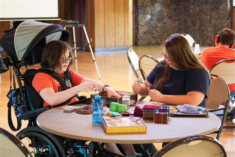 Two people at a round table engaging in arts and crafts, with various materials and supplies spread out. One uses a wheelchair.
