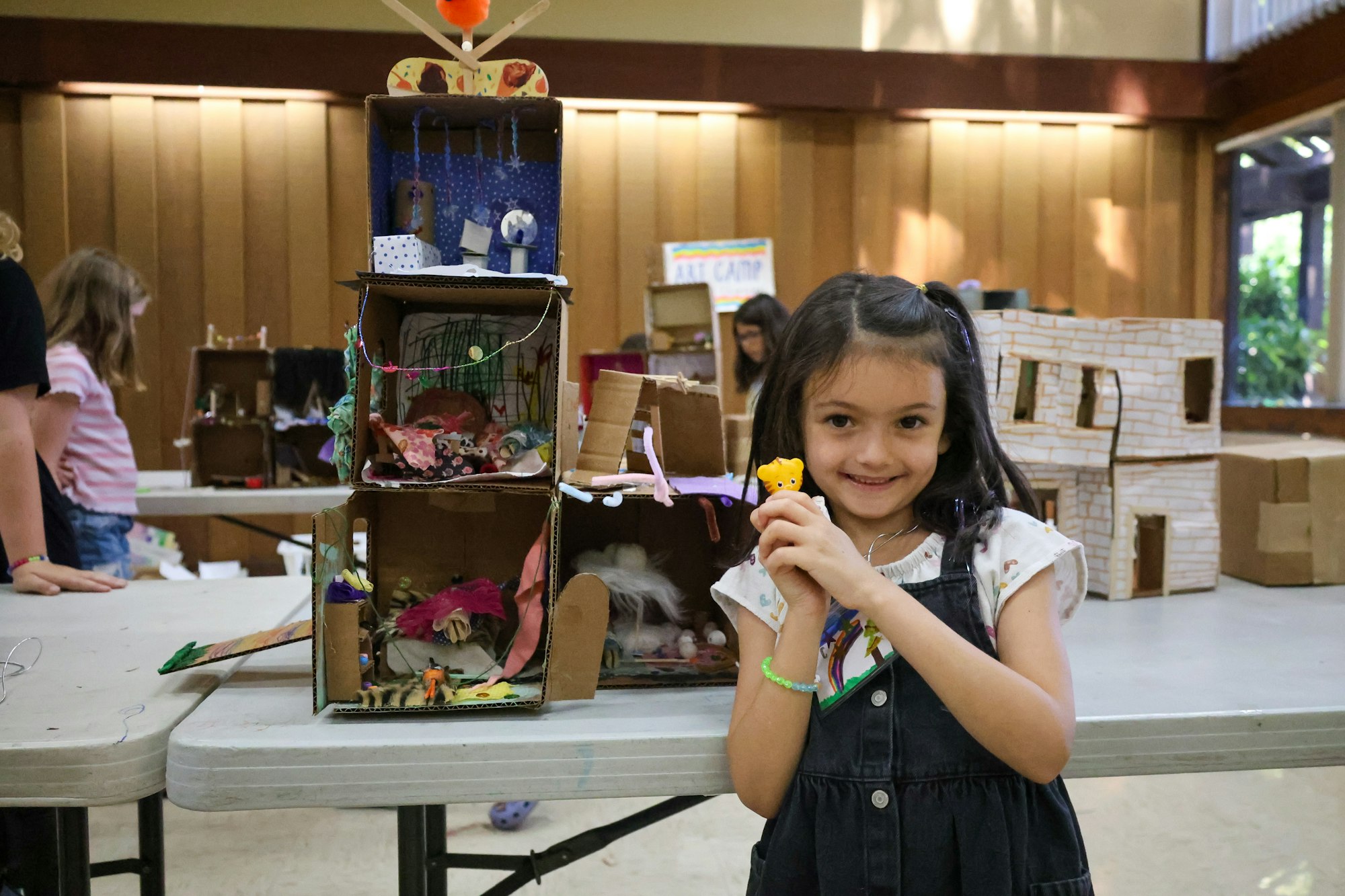 A girl at a crafts table, holding a toy, with homemade cardboard projects around her.