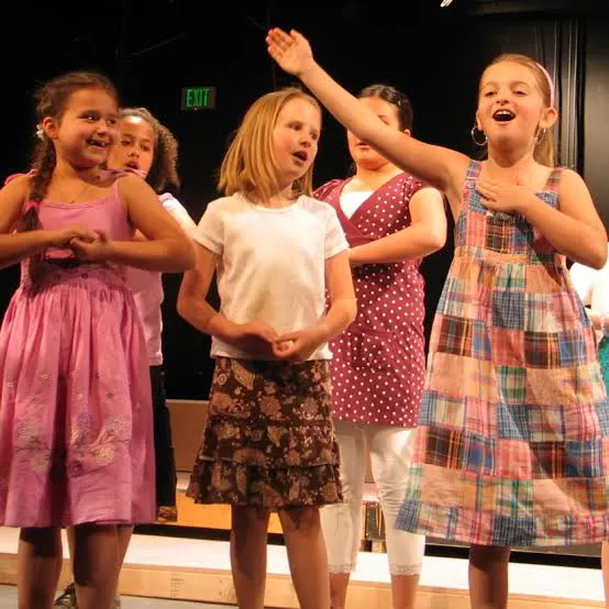 A group of young girls is performing on stage, expressing joy and participating in a lively musical or theatrical act.