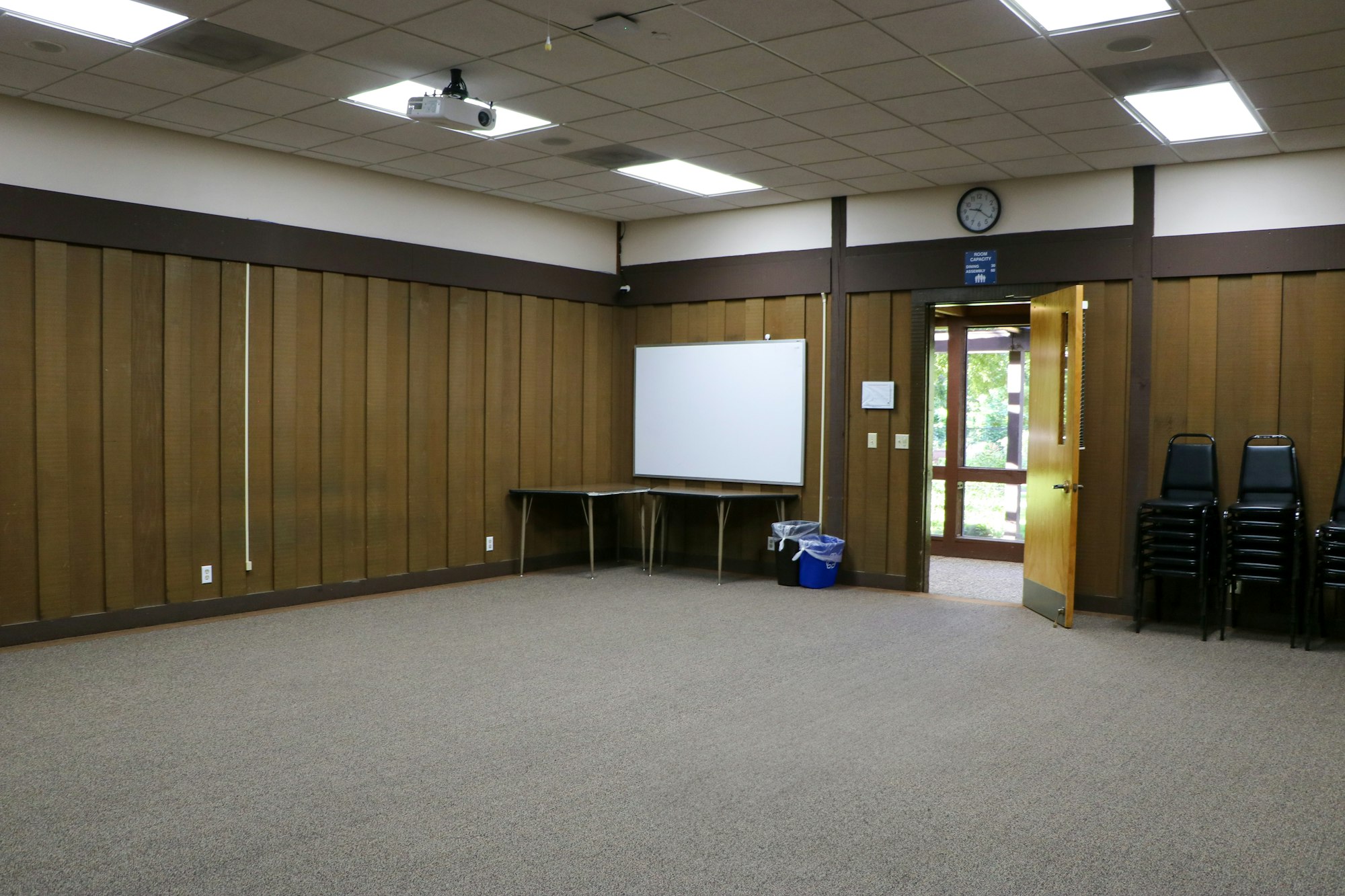 An empty conference room with chairs stacked, a projector, a projection screen, a clock, and a door to the outside.