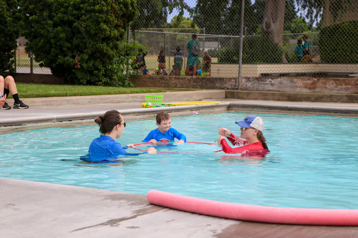 Three people are in a swimming pool, engaging in activities with a child while others watch from the edge. Fun and learning time!