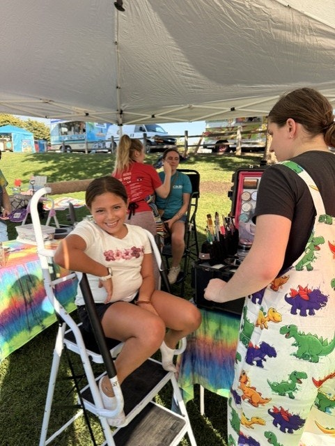 A young girl is seated for face painting at an outdoor event, while others chat nearby at a colorful booth.