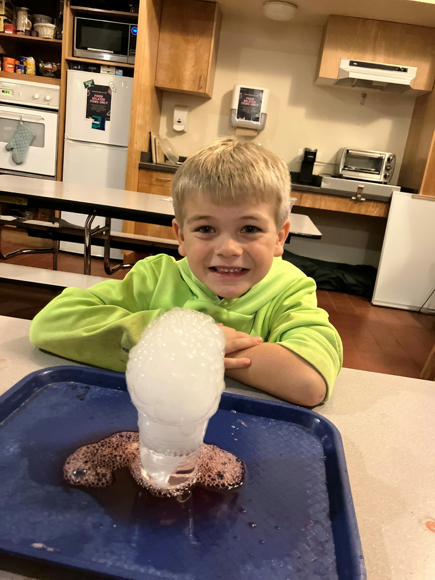 A child in a green hoodie smiling at a bubbling science experiment on a tray in a kitchen setting.
