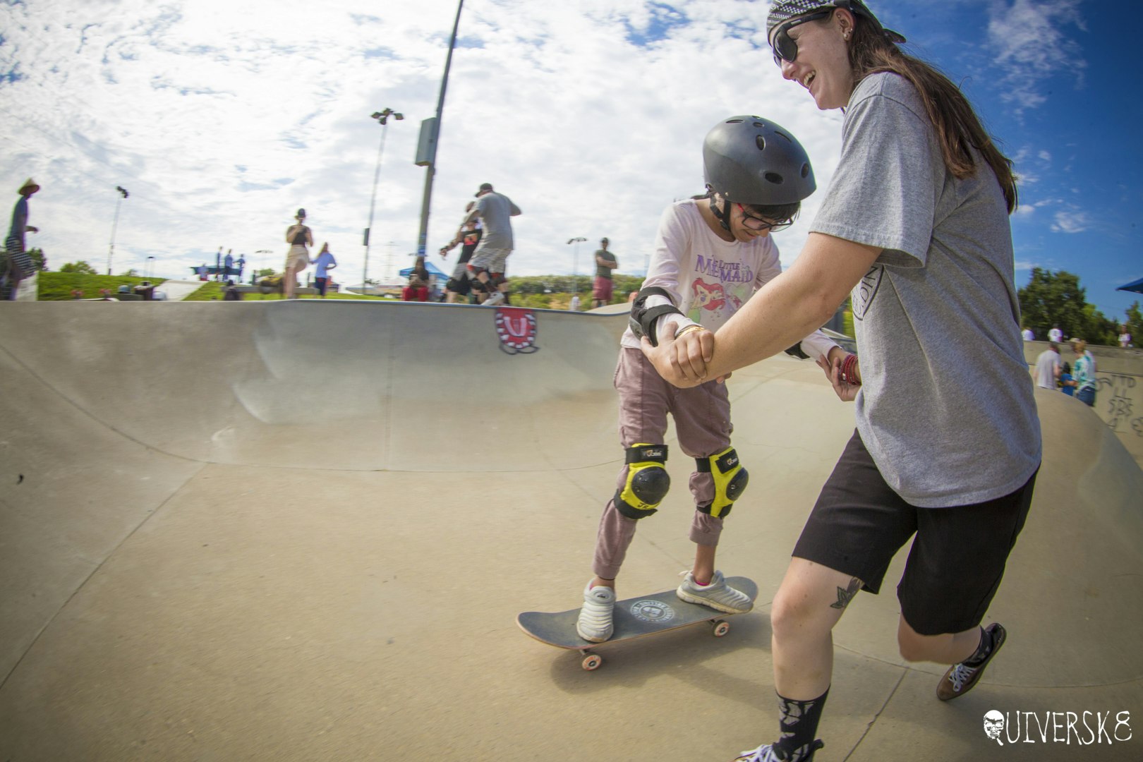 Two people at a skatepark, one learning to skateboard with assistance.