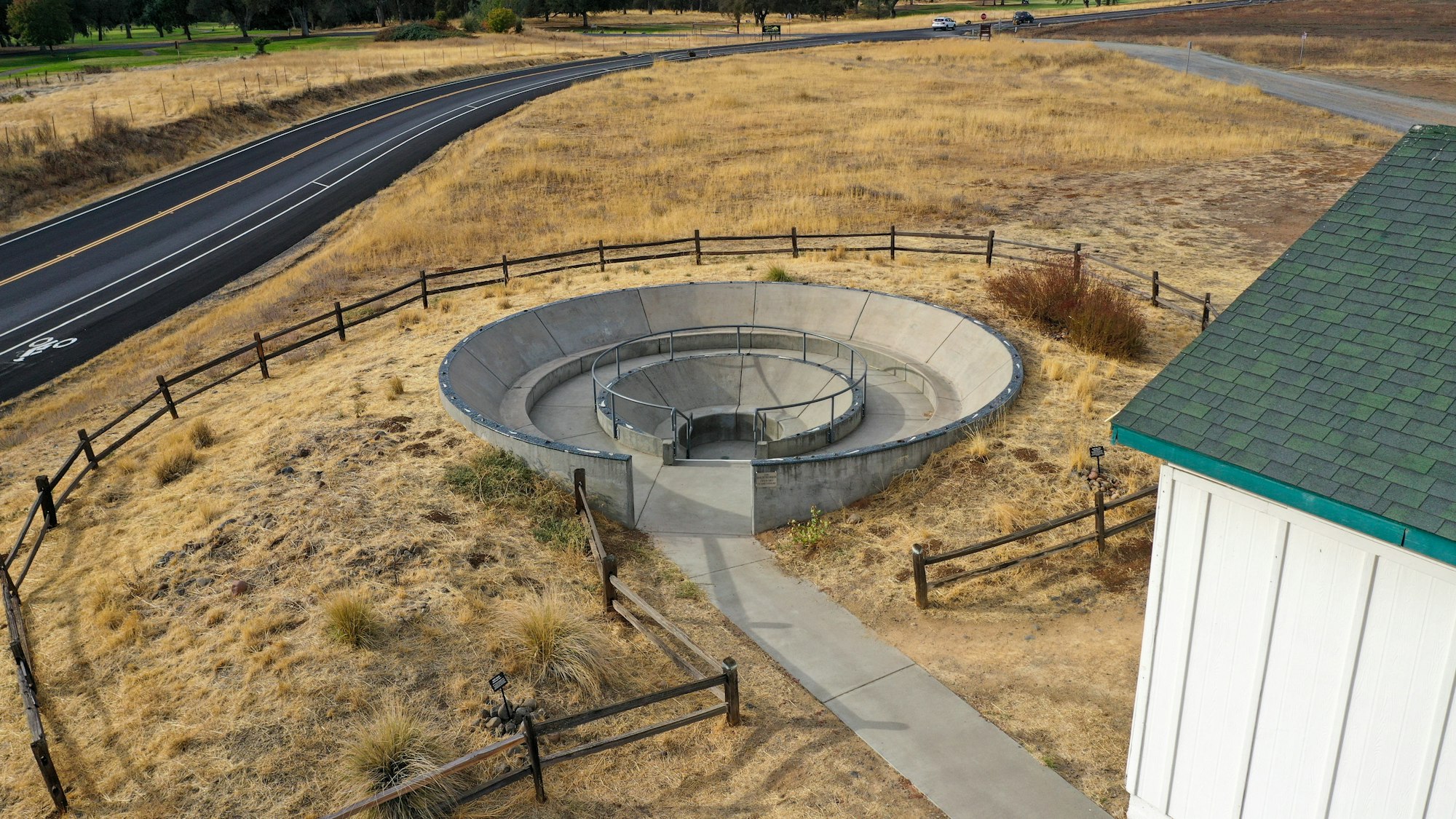 Spiral concrete staircase structure in a grassy area next to a road, surrounded by wooden fencing.
