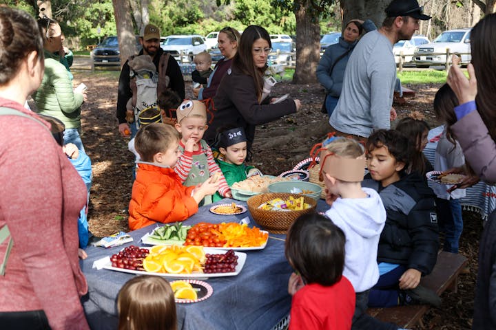 A group of children and adults gather outdoors, enjoying a feast with fruits and snacks, while wearing festive hats.