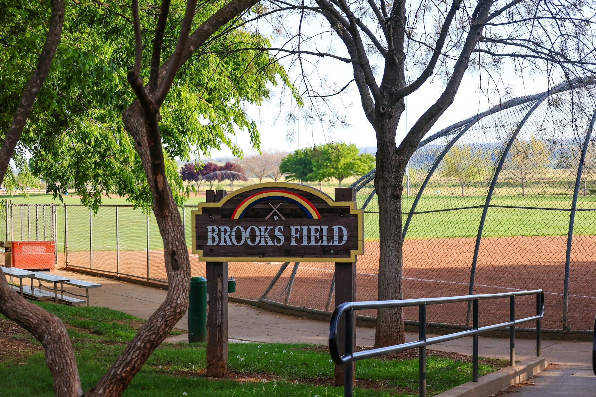 Sign for "Brooks Field" next to a baseball field, with trees and a fence in the background.