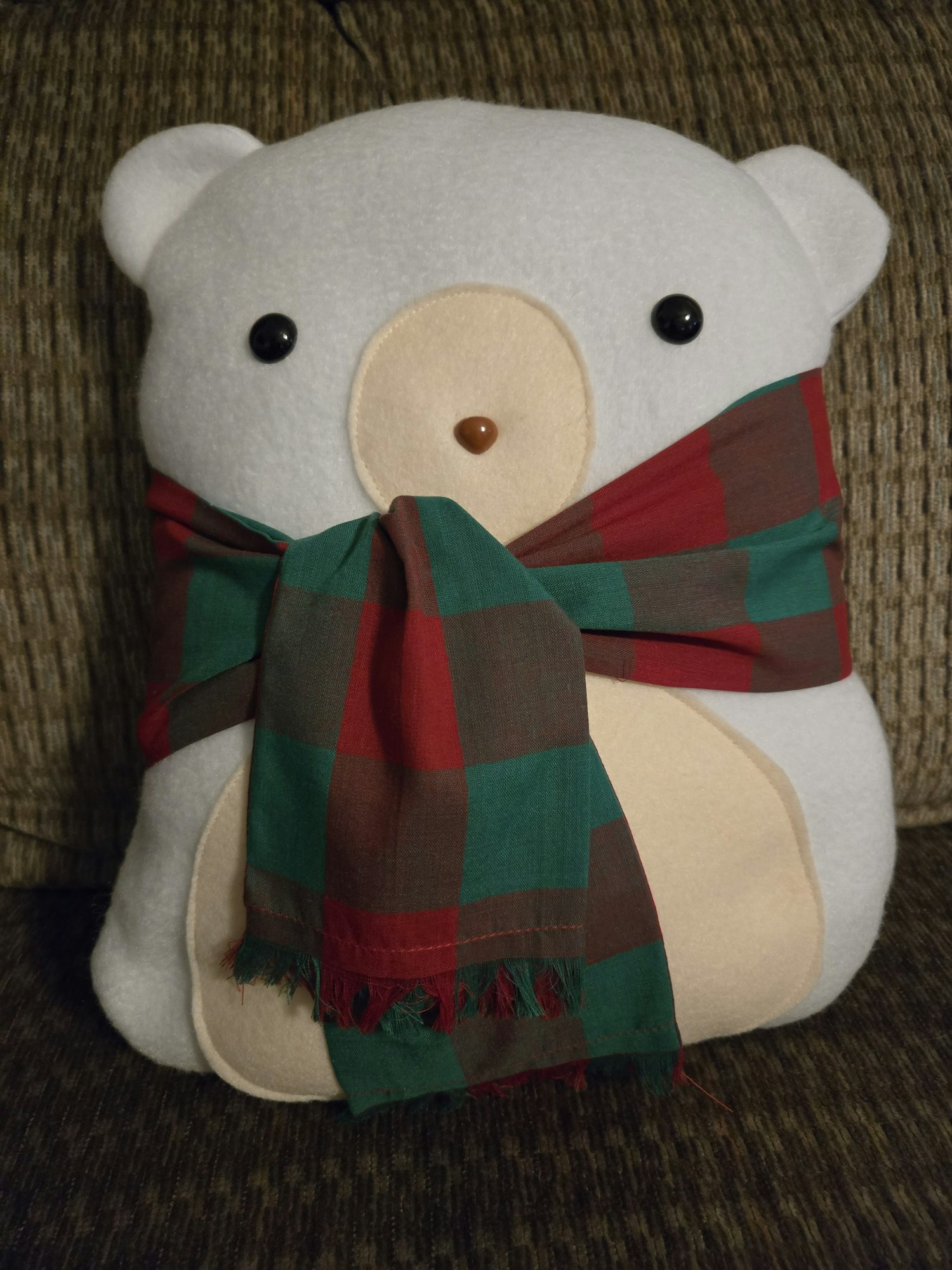 A white bear-shaped cushion wearing a colorful plaid scarf, resting on a textured couch.