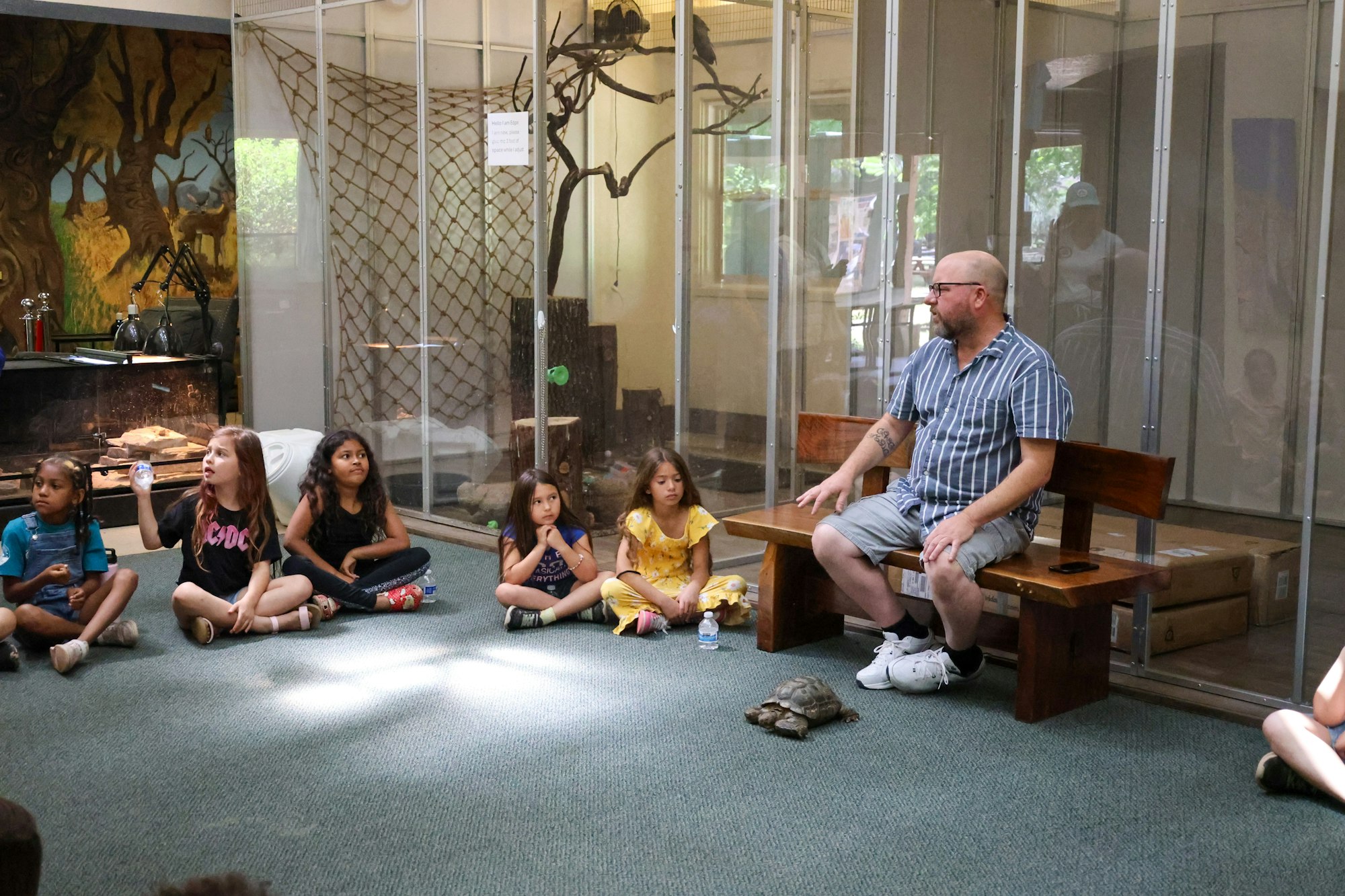 A man and children sit in a circle on the floor with a tortoise in the center, near a glass enclosure.