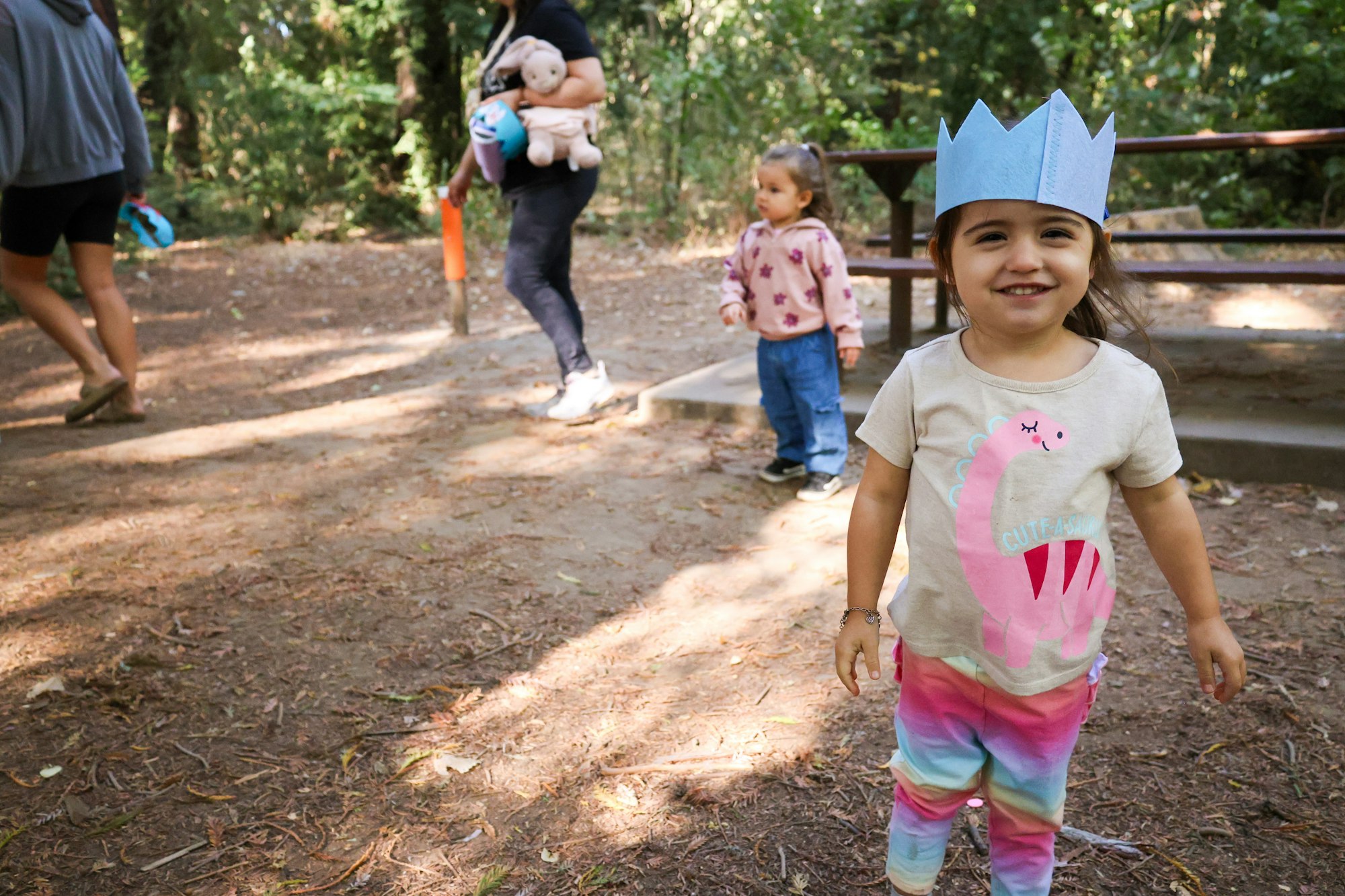 A young girl in a blue paper crown smiles in a wooded area, while others walk nearby. Playful and cheerful scene.