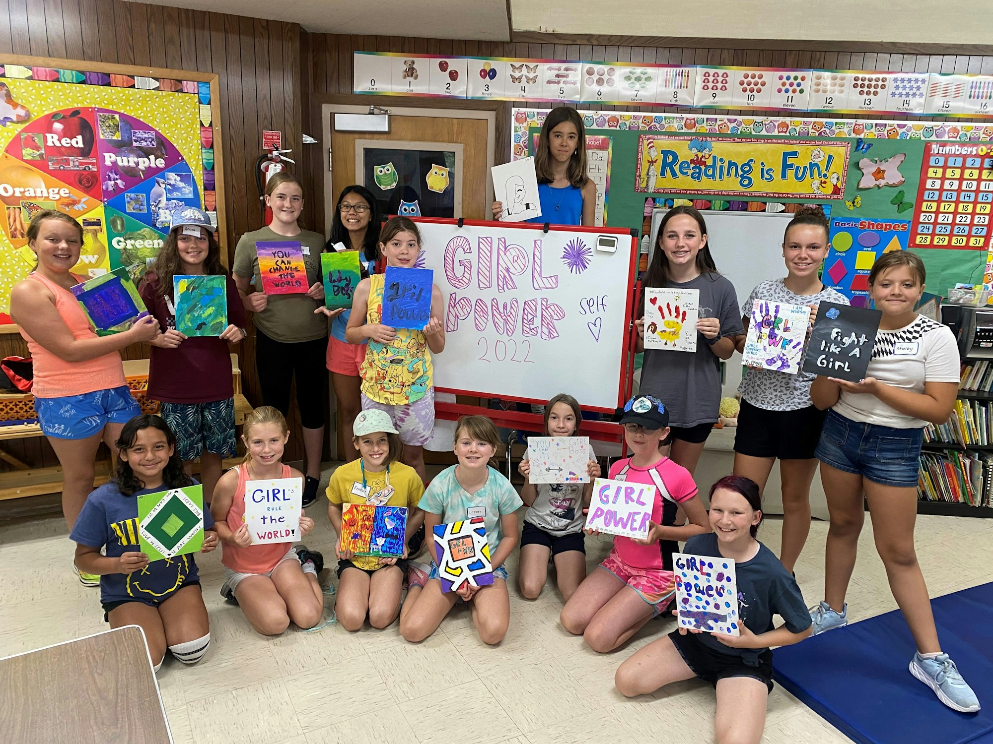 Group of girls holding paintings with empowering messages, standing around a "Girl Power 2022" sign in a classroom.