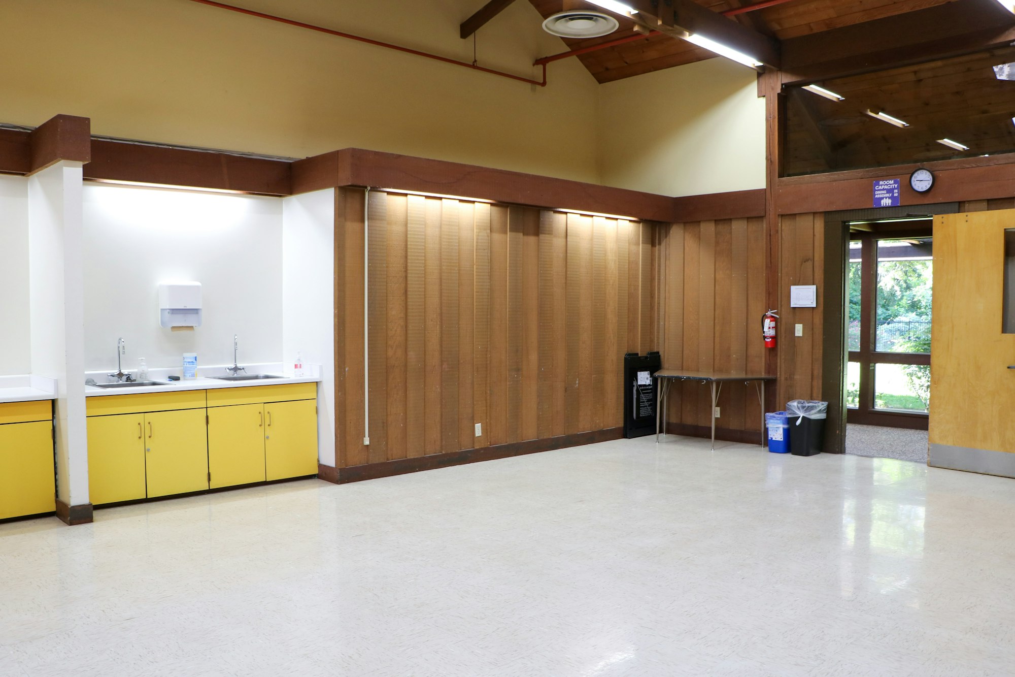 Empty room with wooden partitions, yellow cabinets with sinks, a clock, recycling bins, and a glass door leading outside.