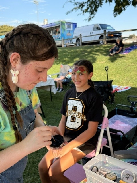 At CARD Movies in the park a young girl is getting her face painted at an outdoor event, with food trucks and people in the background.