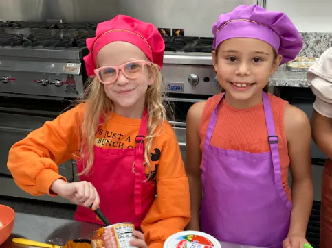 Two children in colorful chef hats are smiling while preparing food in a kitchen. One is stirring a bowl.