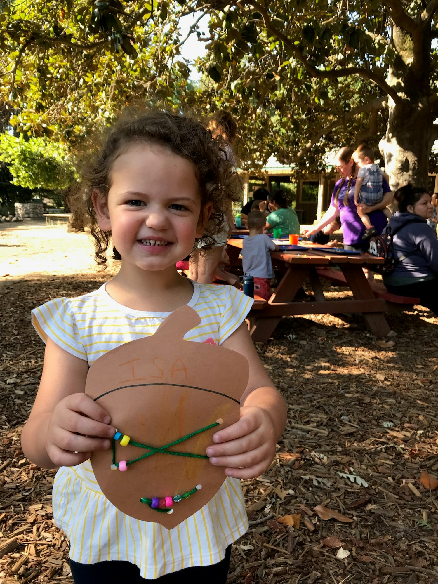 A smiling girl holds a decorated acorn craft with beads, surrounded by a park setting with other children and adults in the background.