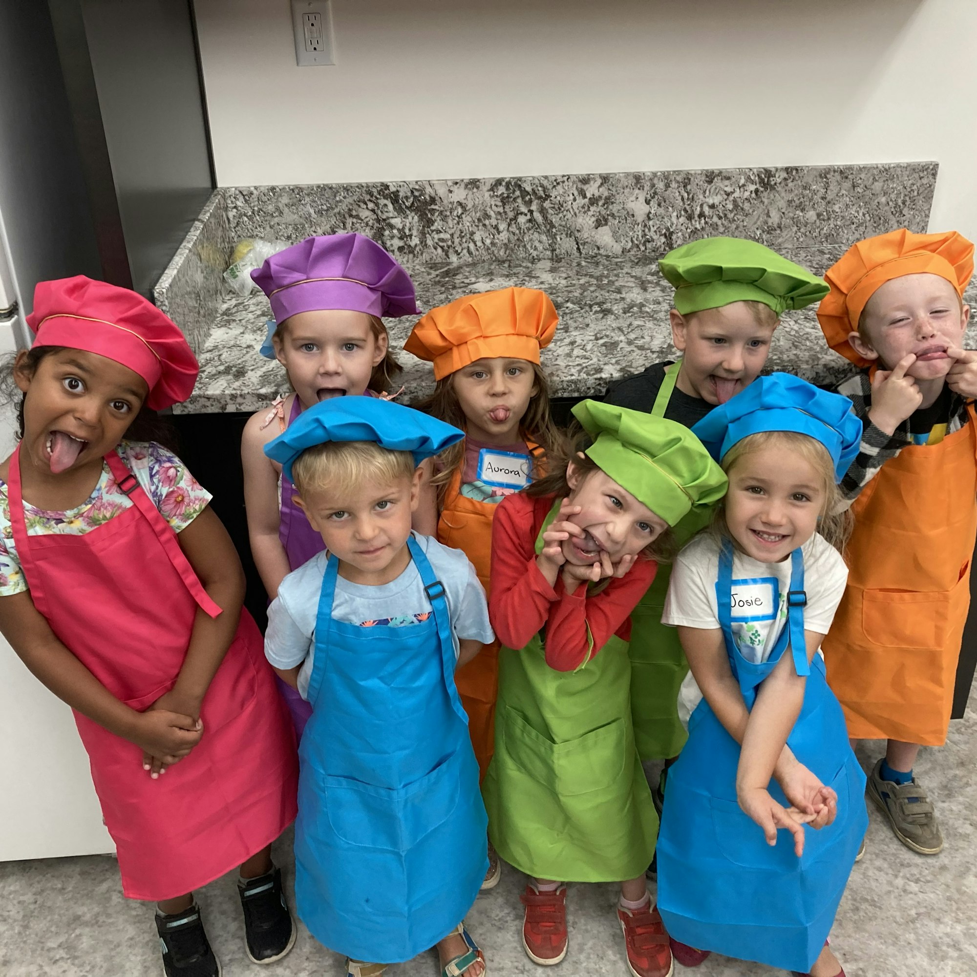 A group of kids in colorful chef hats and aprons making funny faces, posing together in a kitchen setting.
