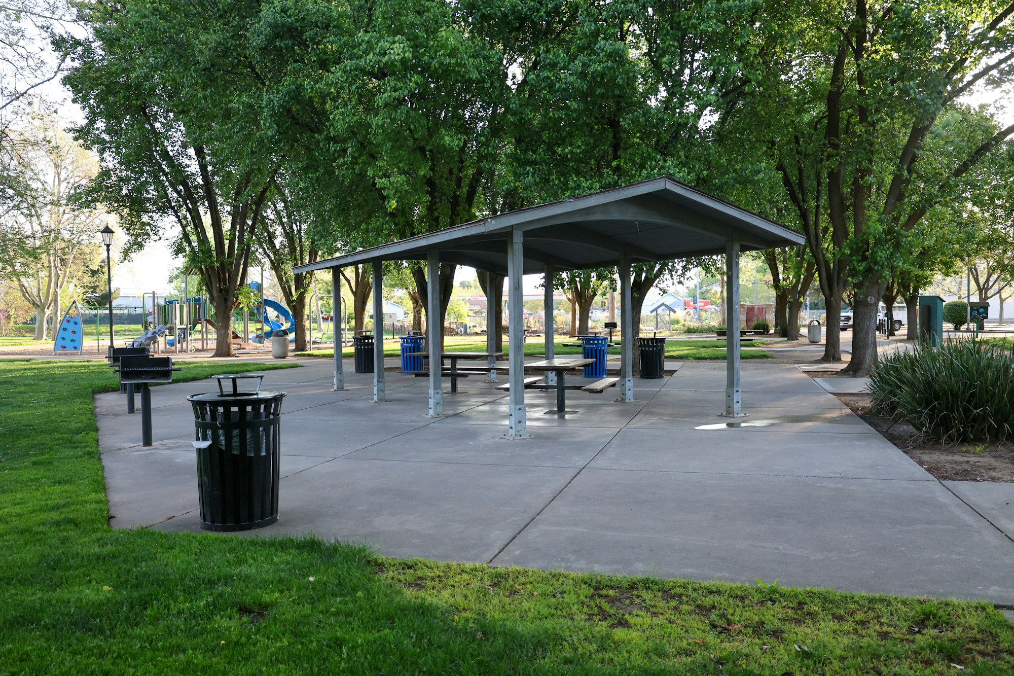 A park with a picnic shelter, benches, trash cans, and a playground in the background, surrounded by lush trees.
