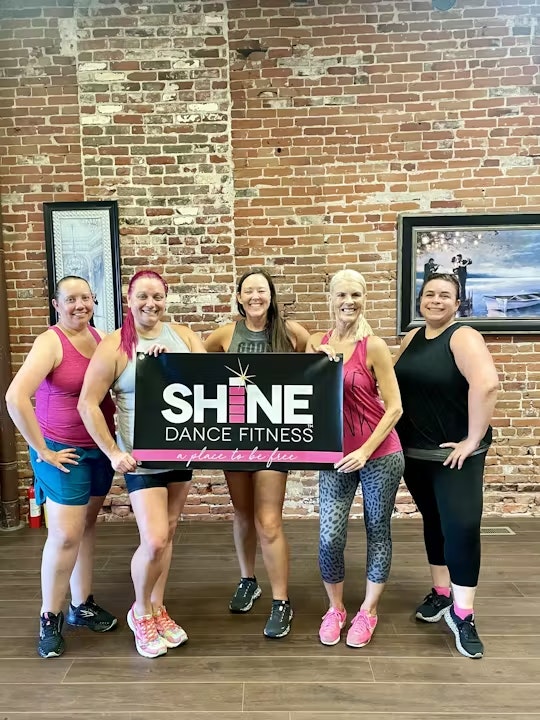 A group of five women holding a "SHINE Dance Fitness" sign in a fitness studio with a brick wall background.