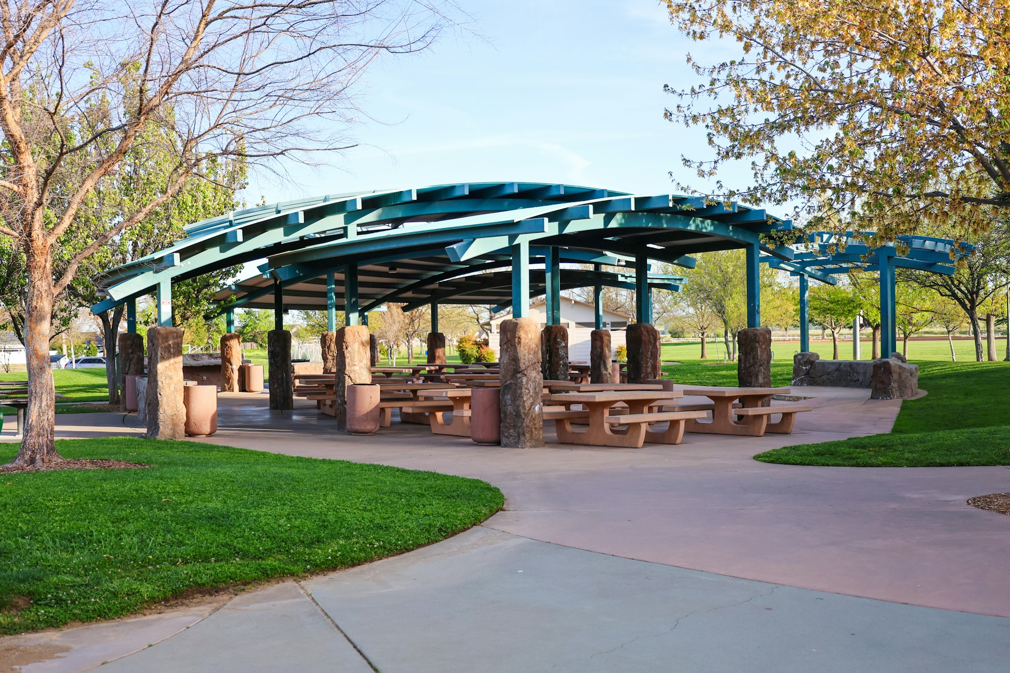 A park shelter with picnic tables, green grass, trees, and a walking path under a clear sky.