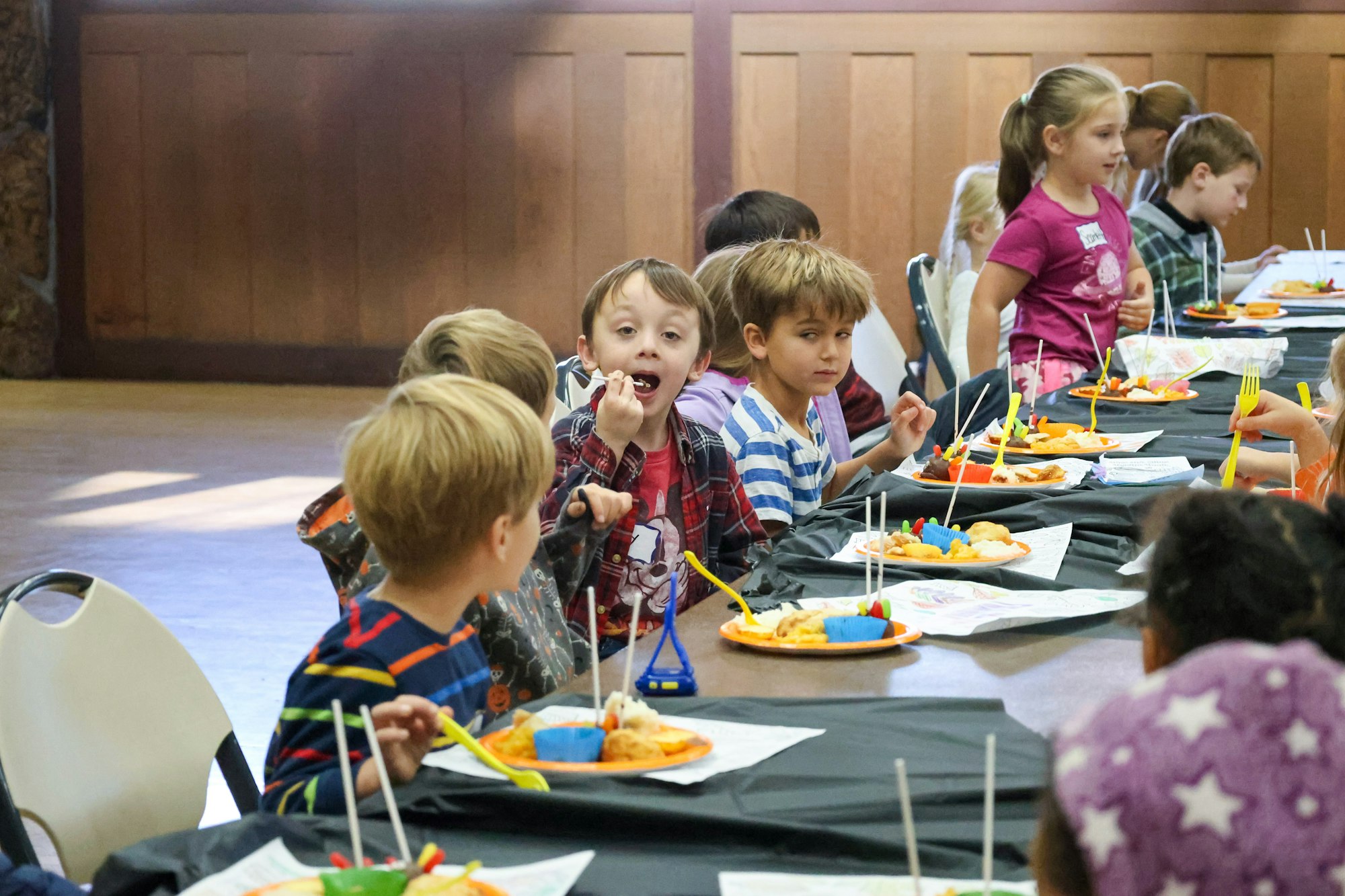 Children sitting at a table eating, with plates of food and drinks, in a social gathering or party setting.