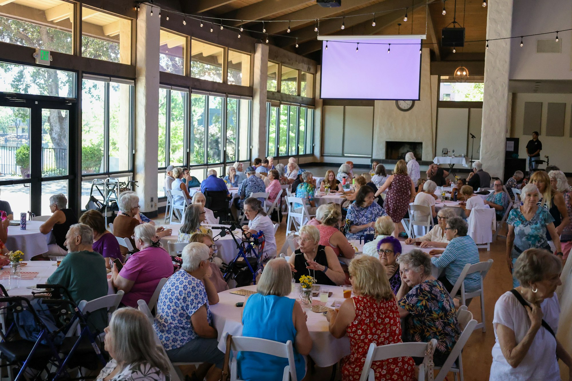 A large gathering of people, mostly seniors, seated at tables in a bright indoor space with large windows and a screen.