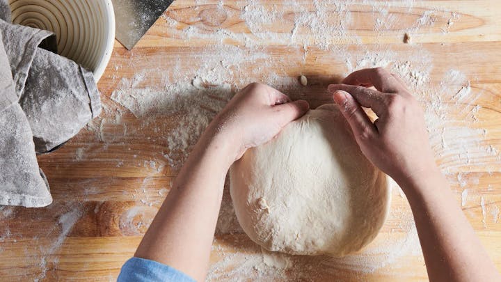 Hands shaping dough on a floured wooden surface, with a dough scraper and banneton nearby.