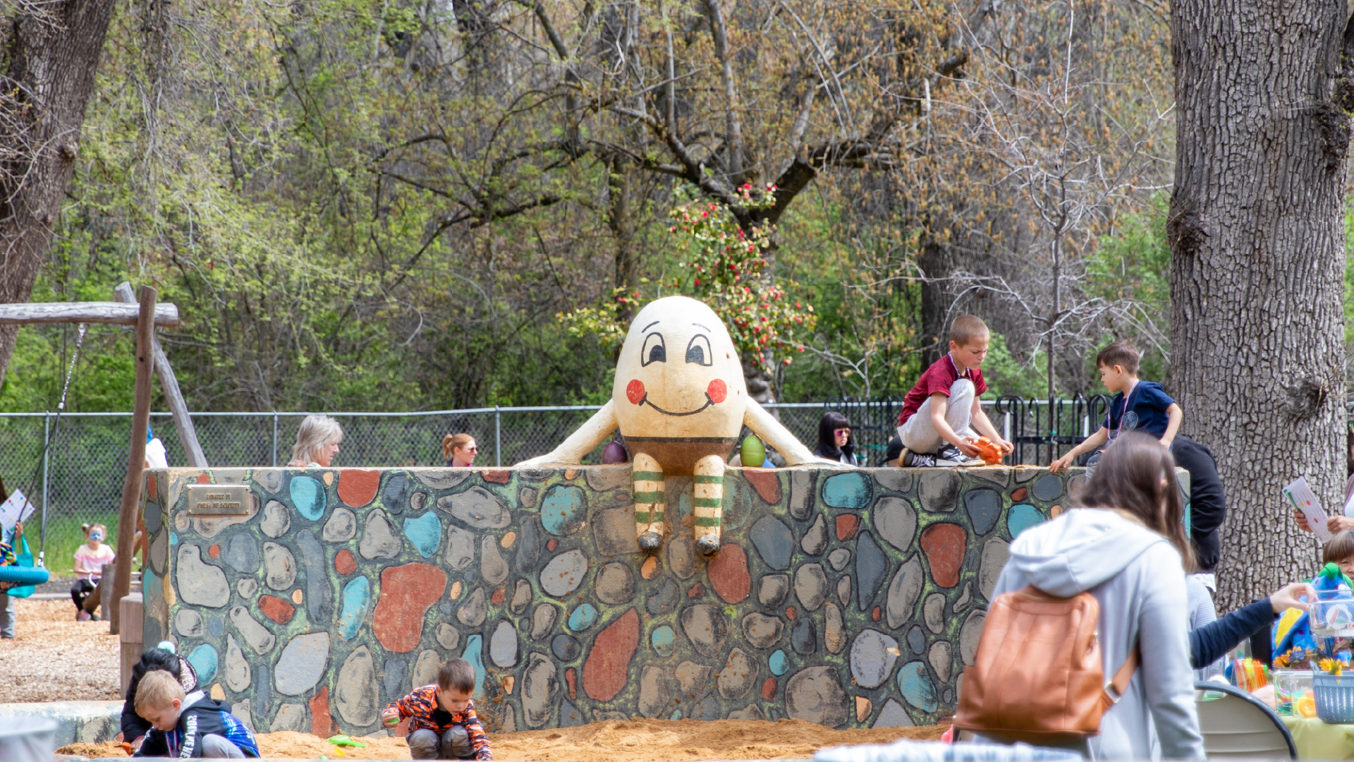 A playground with children, a large egg-shaped character sculpture, trees, and some adults.