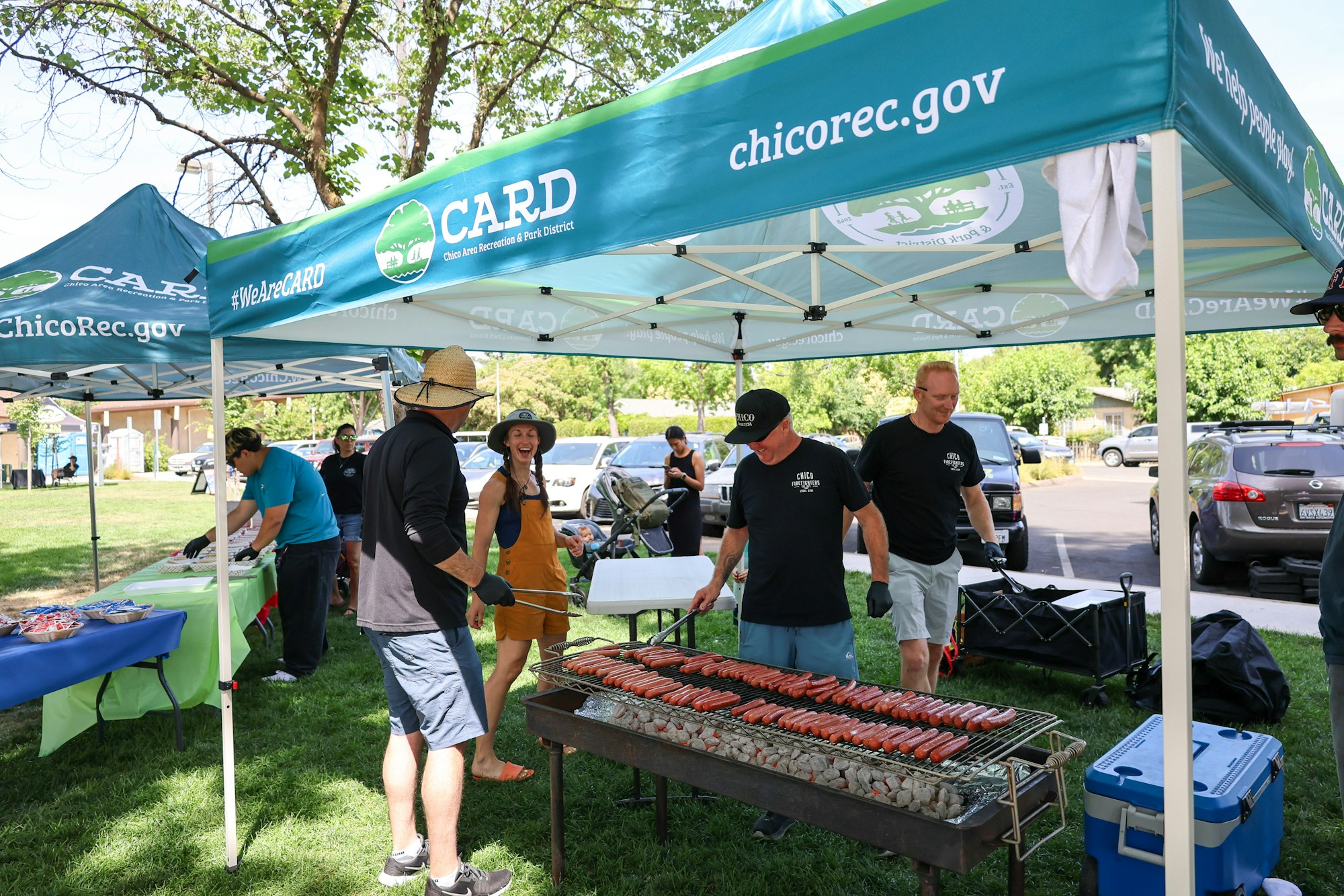 A group is cooking hot dogs at a community event under tents promoting the Chico Area Recreation and Park District.
