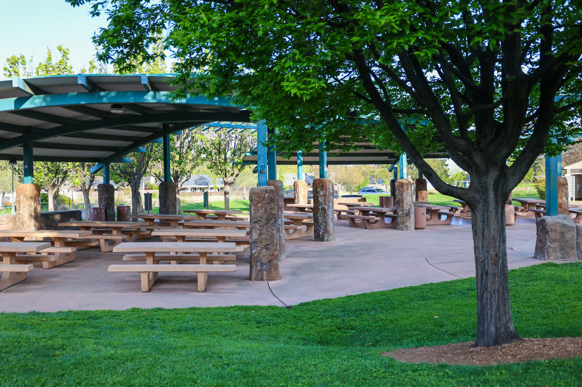 A park with picnic tables, trees, and a covered pavilion on a sunny day.