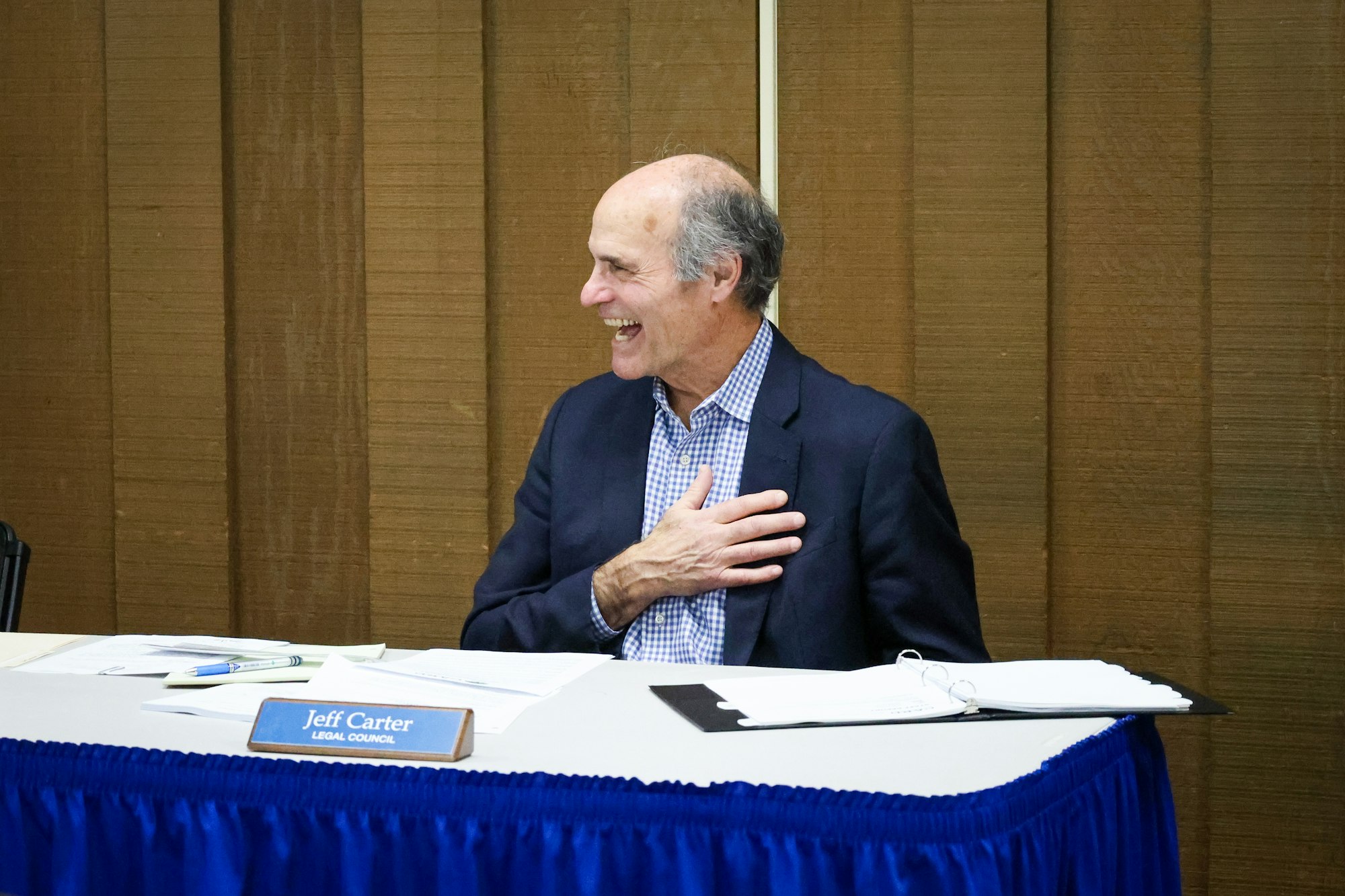 A smiling man seated at a table with a nameplate that reads "Legal Council" and papers in front of him.